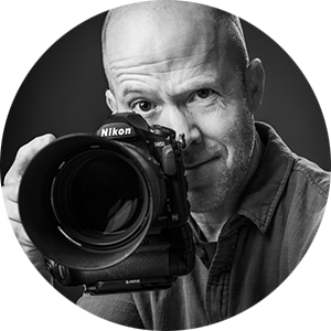 B&W image of photographer Matt Kosterman with a raised eyebrow holding one of his Nikon D850 cameras up